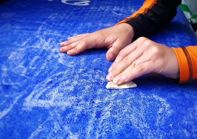 A person's hands kneading a blue clay bar into a flattened shape, ready for use on a car. The background is blurred, showing detailing supplies like spray bottles and microfiber cloths.