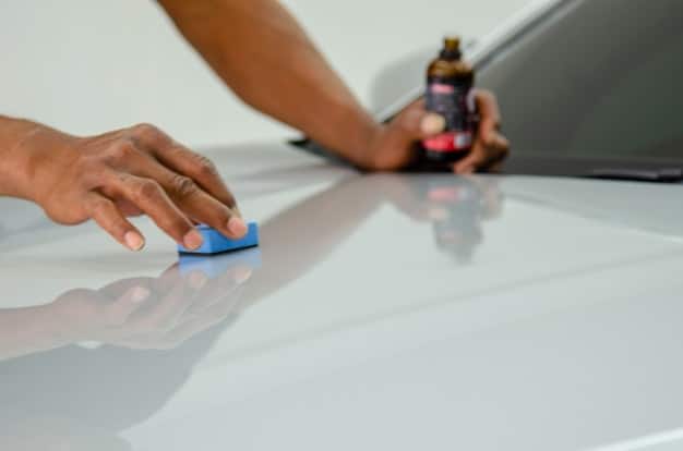 A hand applying a thin, even layer of wax onto the polished surface of a car, with the reflection of the surrounding environment visible on the paint. The focus is on the application technique and the protective quality of the wax.