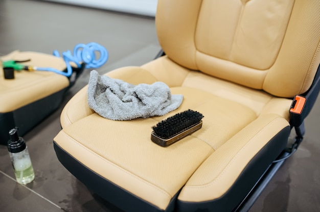 A collection of leather cleaning supplies arranged on a car seat. The set includes a bottle of leather cleaner, a bottle of leather conditioner, a soft microfiber towel, and a soft-bristled brush. The scene is brightly lit to showcase the products clearly.