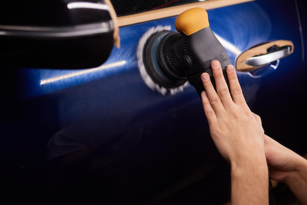 A car detailing professional using a power tool to polish a headlight. The surrounding area is taped off to protect the car's paint.