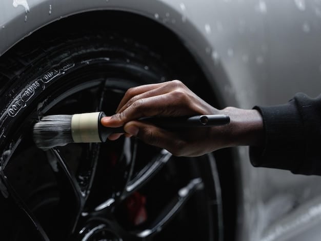 Close-up of a person using a tire brush to scrub the sidewall of a car tire. The brush is agitating a foamy cleaning product, and grime is visibly being lifted from the rubber. The focus is on the cleaning action and the tools used in tire detailing.