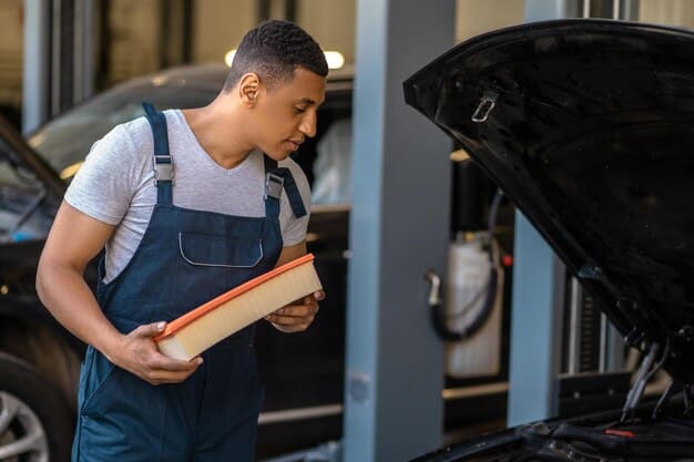 A mechanic pointing to the air filter housing in an engine bay, explaining the location and how to open it. The mechanic is wearing gloves and using a tool to demonstrate. DIY Air Filter Replacement: Improve Fuel Efficiency by Up to 10% should be mentioned in the photo.