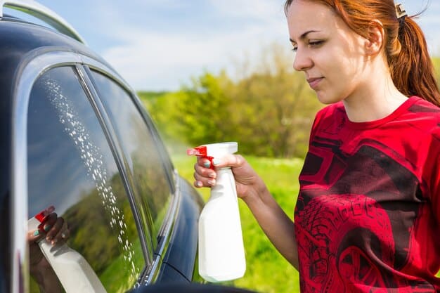 A person washing a car using a waterless car wash product in a spray bottle. The car is parked on a paved surface and the background shows a residential neighborhood, emphasizing the ease and convenience of waterless washing.