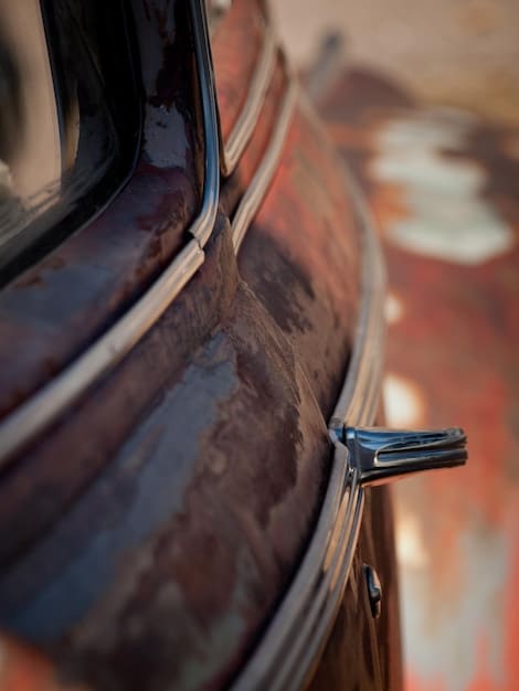 A close up of a car's undercarriage being treated with a rust-proofing spray during a winter car detailing session. The focus is on the nozzle of the spray can and the area being coated. The technician is wearing gloves and protective eyewear. The surroundings show a well-lit garage with professional detailing equipment.